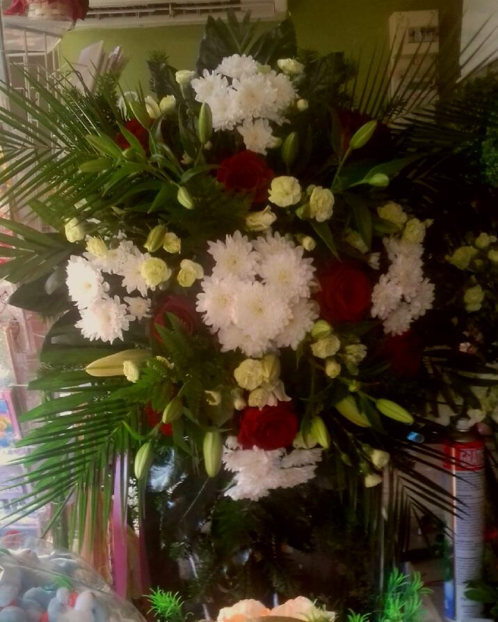 Duża kompozycja kwiatowa w szklanym wazonie: białe chryzantemy, czerwone róże, zielone liście palmowe i białe lilie.