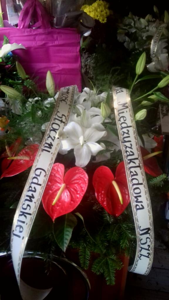 Wieniec pogrzebowy z czerwonymi anturiami i białymi liliami, z szarfami z napisem 'Stoczni Gdańskiej' i 'Międzyzakładowa N572', na tle ciemnego otoczenia.