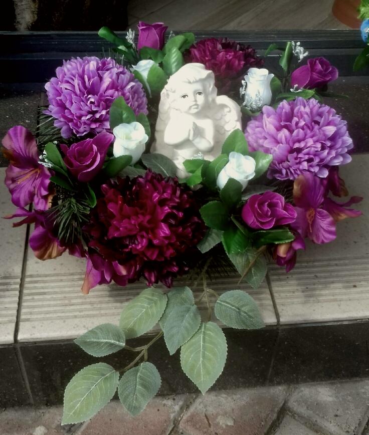 Kompozycja nagrobna z fioletowych i bordowych kwiatów oraz białej figurki anioła z złożonymi rękami, ułożona na płytkach.