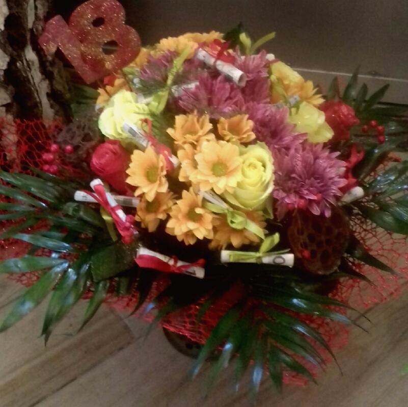 Urodzinowy bukiet z żółtych, pomarańczowych i fioletowych kwiatów, udekorowany rulonikami z banknotami przewiązanymi wstążkami oraz czerwoną siatką i brokatową liczbą 18, stojący obok pnia drzewa.