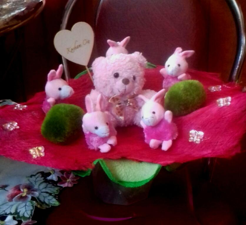 Różowy pluszowy miś otoczony małymi króliczkami i zielonymi kulkami na czerwonym, futrzanym podłożu, ozdobionym błyszczącymi motylkami, z sercem z napisem 'Kocham Cię'.