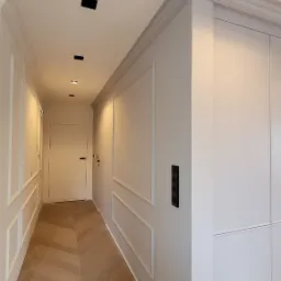 Nowoczesny korytarz z jasnymi ścianami z dekoracyjnymi listwami, parkietem ułożonym w jodełkę i minimalistycznym oświetleniem punktowym.