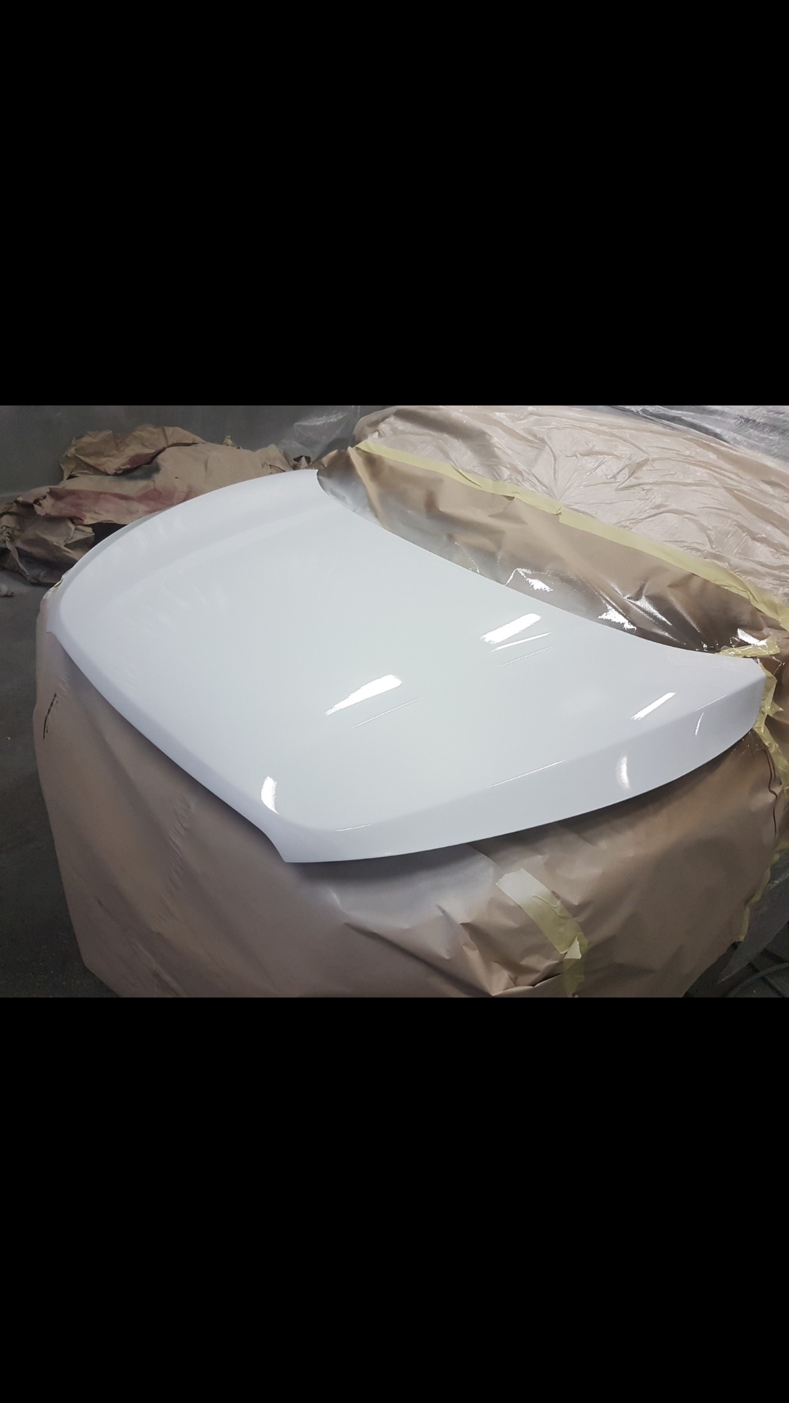 Pomalowana na biało maska samochodu, leżąca na papierze maskującym, widoczne odbicia światła na świeżej powłoce lakierniczej.