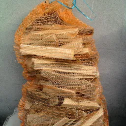 Worek z pomarańczowej siatki wypełniony pociętymi kawałkami drewna na jasnym tle, zawiązany niebieskim sznurkiem.