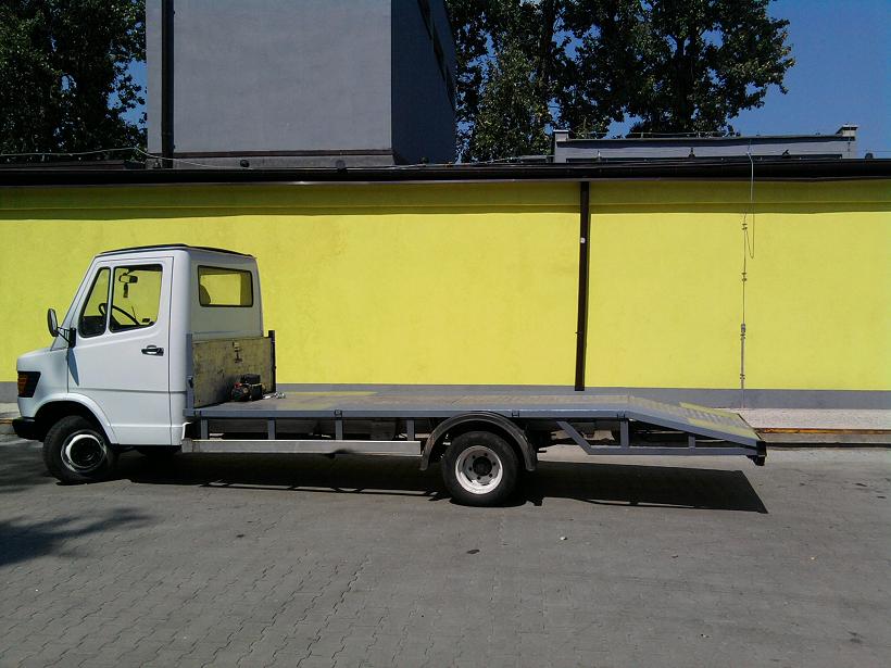 Biała laweta z platformą transportową na tle jaskrawożółtej ściany z czarnymi elementami pionowymi.