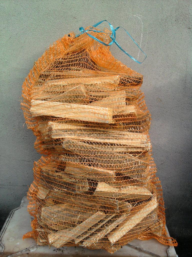 Worek z pomarańczowej siatki wypełniony pociętymi kawałkami drewna na jasnym tle, zawiązany niebieskim sznurkiem.