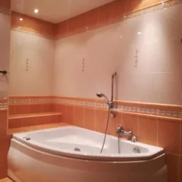 Jasna łazienka z narożną wanną z hydromasażem, obłożona glazurą w odcieniach beżu i pomarańczy. Oświetlenie punktowe sufitowe. Bateria wannowa chromowana.