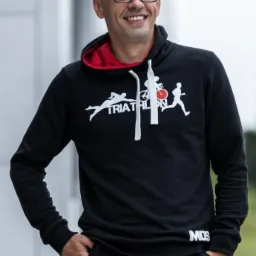 Uśmiechnięty mężczyzna w okularach, ubrany w czarną bluzę z kapturem z grafiką triathlonu, pozuje na zewnątrz. Widoczny napis MOS na bluzie.