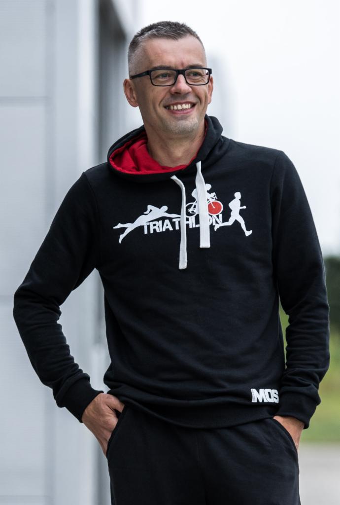 Uśmiechnięty mężczyzna w okularach, ubrany w czarną bluzę z kapturem z grafiką triathlonu, pozuje na zewnątrz. Widoczny napis MOS na bluzie.