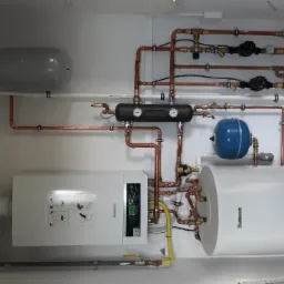 Kompletna instalacja centralnego ogrzewania z kotłem gazowym Buderus, zasobnikiem ciepłej wody, naczyniami wyrównawczymi i rozdzielaczem, widoczne miedziane rury i osprzęt.