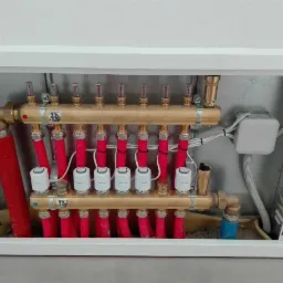 Rozdzielacz ogrzewania podłogowego w białej szafce, z czerwonymi rurkami i białymi głowicami termostatycznymi, widoczna skrzynka elektryczna z białymi kablami.