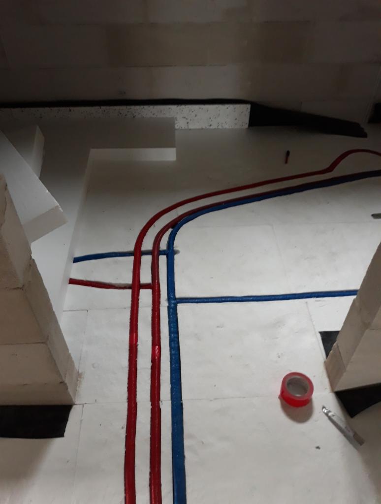 Instalacja hydrauliczna: rury w kolorach czerwonym i niebieskim ułożone na białej izolacji podłogowej, widoczna taśma izolacyjna i nóż introligatorski.