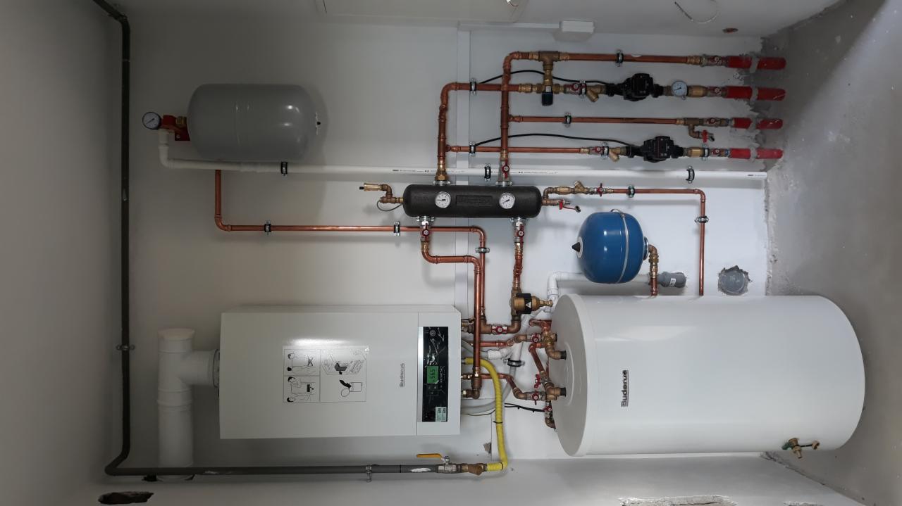 Kompletna instalacja centralnego ogrzewania z kotłem gazowym Buderus, zasobnikiem ciepłej wody, naczyniami wyrównawczymi i rozdzielaczem, widoczne miedziane rury i osprzęt.
