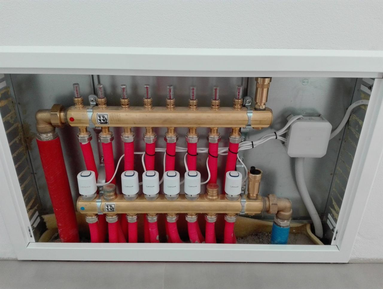 Rozdzielacz ogrzewania podłogowego w białej szafce, z czerwonymi rurkami i białymi głowicami termostatycznymi, widoczna skrzynka elektryczna z białymi kablami.