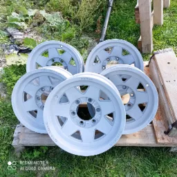 Zestaw pięciu odnowionych, szarych felg aluminiowych do samochodów terenowych, leżących na drewnianej palecie na trawie. Na felgach widoczne napisy 'OFF ROAD' i 'melber'.