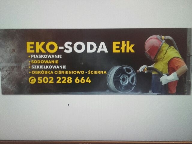 Baner reklamowy firmy Eko-Soda Ełk, oferującej obróbkę strumieniowo-ścierną felg. Na zdjęciu pracownik w kombinezonie ochronnym czyści felgę.
