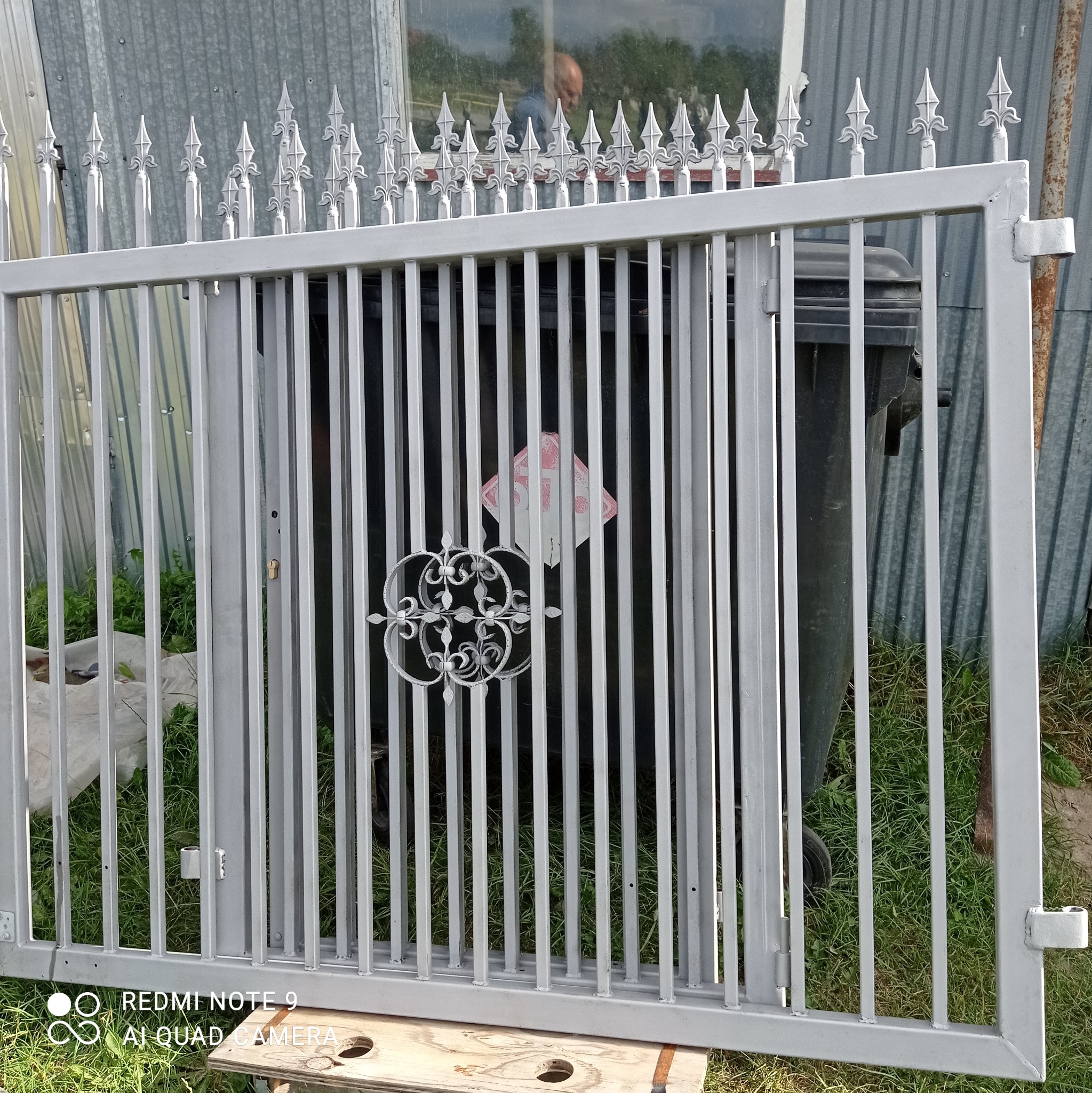 Szara, metalowa brama z dekoracyjnymi grotami i ozdobnym elementem pośrodku, leżąca na drewnianym podeście przed blaszakiem i śmietnikiem.