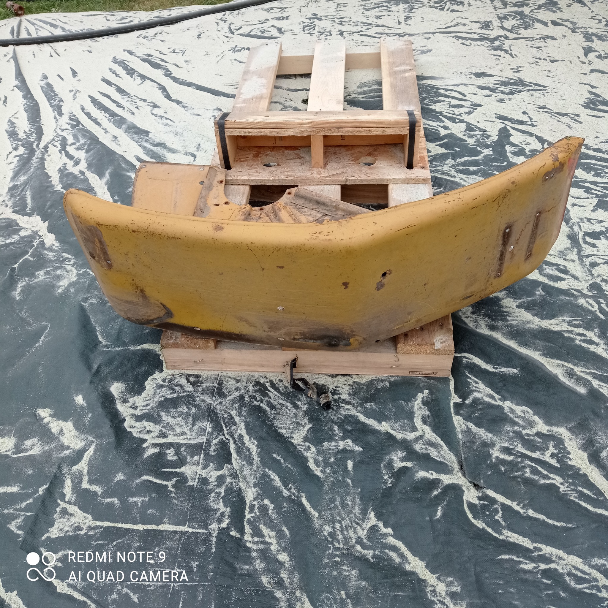 Żółty element karoserii pojazdu, prawdopodobnie błotnik, umieszczony na drewnianej palecie, widoczne ślady korozji i zarysowania, tło stanowi szara plandeka z osadem po piaskowaniu.