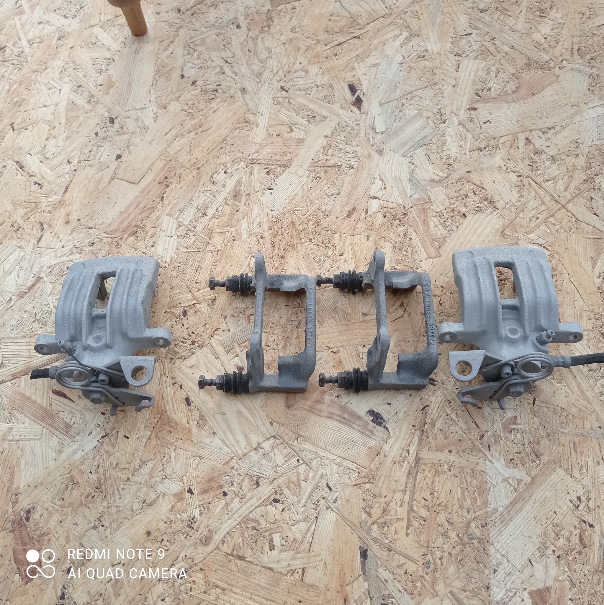 Cztery szare zaciski hamulcowe, dwa kompletne i dwa mocowania, ułożone na płycie OSB.