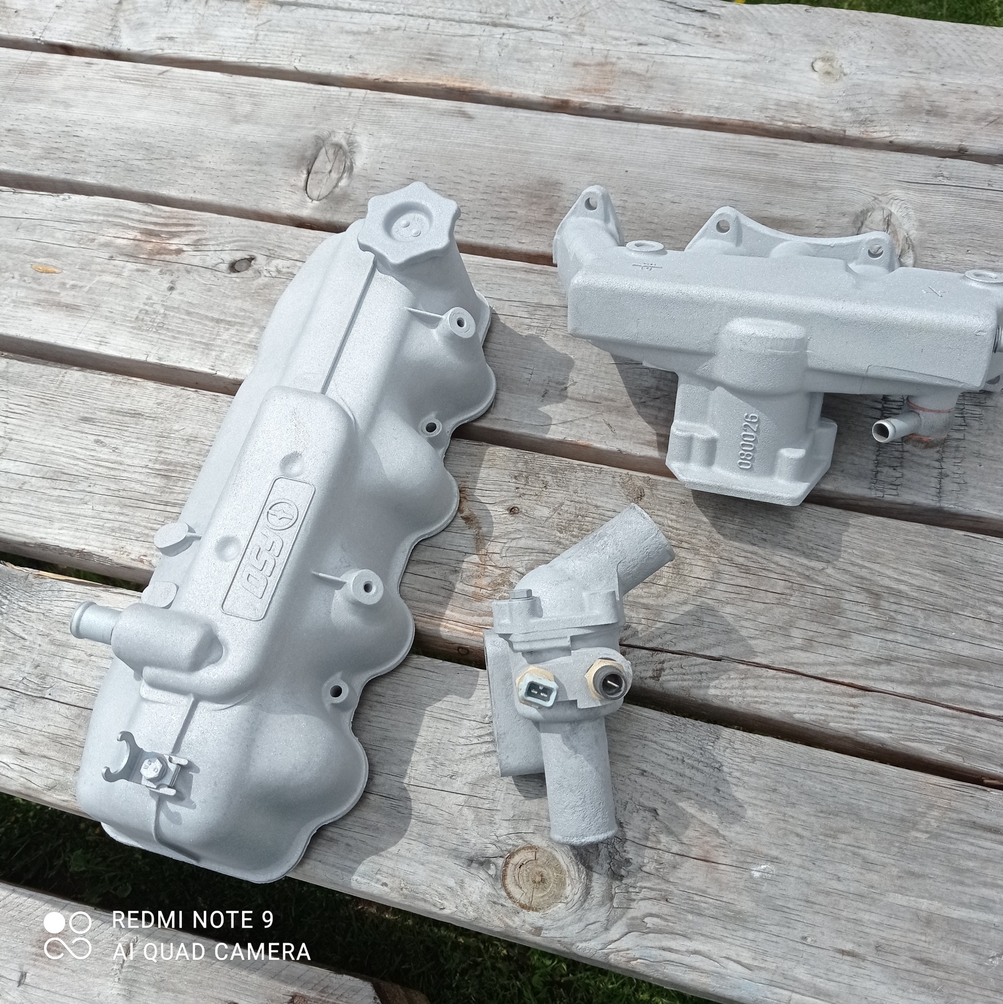 Trzy metalowe elementy samochodowe, pokryte szarą farbą proszkową, leżą na szarym, drewnianym blacie w słoneczny dzień.