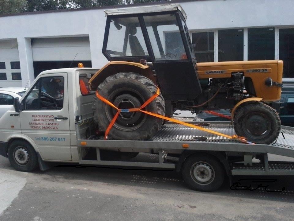 Żółty traktor Ursus C-360 3P zabezpieczony pomarańczowymi pasami na platformie lawety, widoczny napis na lawecie: blacharstwo, lakiernictwo, spawanie plastiku, pomoc drogowa.