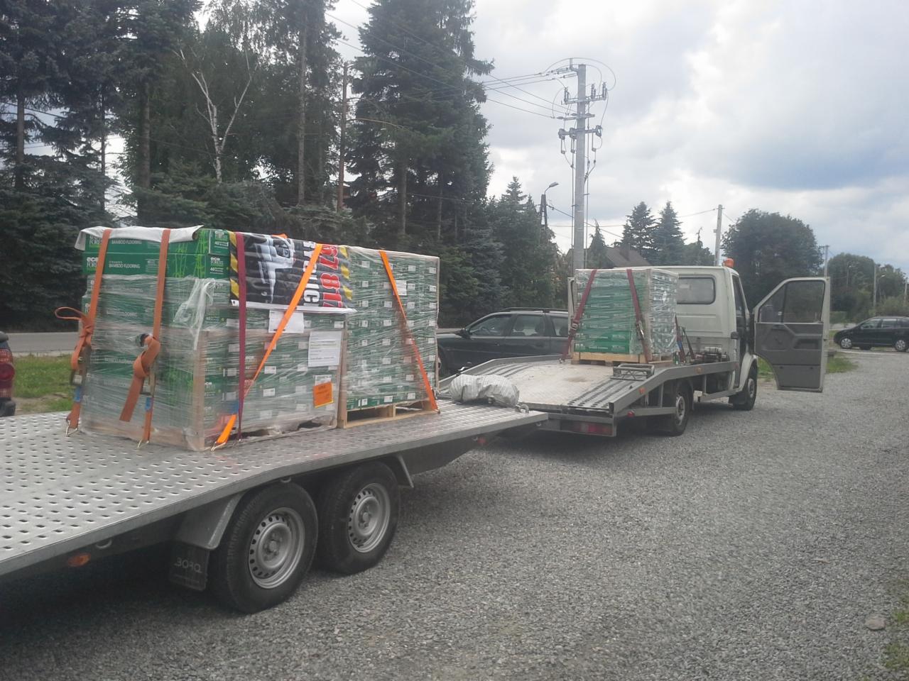 Laweta przewozi dwa palety z zapakowaną podłogą bambusową, zabezpieczone pomarańczowymi pasami transportowymi, widok na otwartą kabinę lawety i otoczenie z drzewami i liniami energetycznymi.