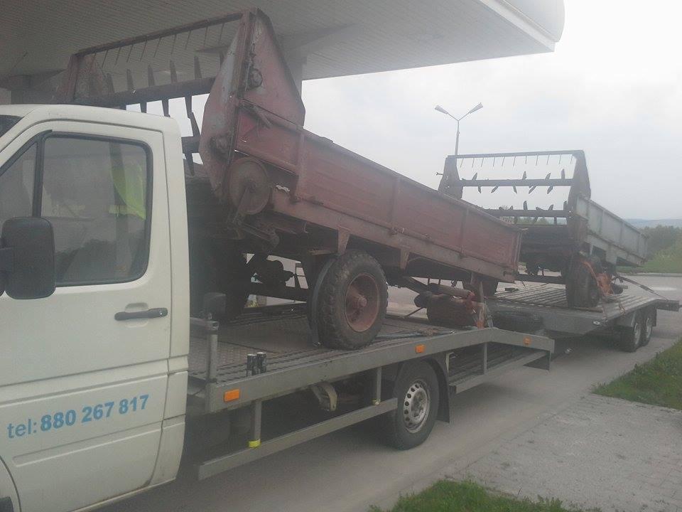 Laweta przewozi dwa stare, rolnicze przyczepy na platformie, widoczne pod światłami stacji benzynowej.