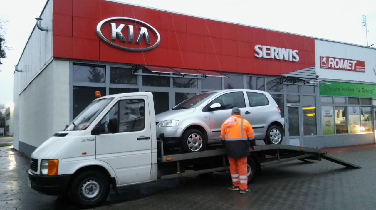 Srebrny samochód osobowy przewożony na lawecie przed budynkiem serwisu samochodowego Kia, pracownik w pomarańczowym kombinezonie zabezpiecza ładunek.