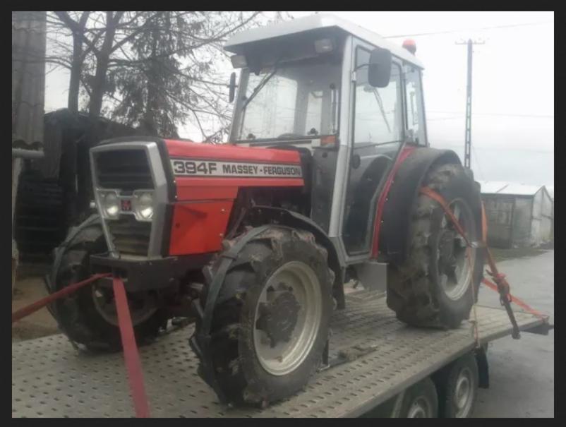 Czerwono-szary traktor Massey Ferguson 394F zabezpieczony pasami na platformie lawety, widok z boku na tle pochmurnego dnia.