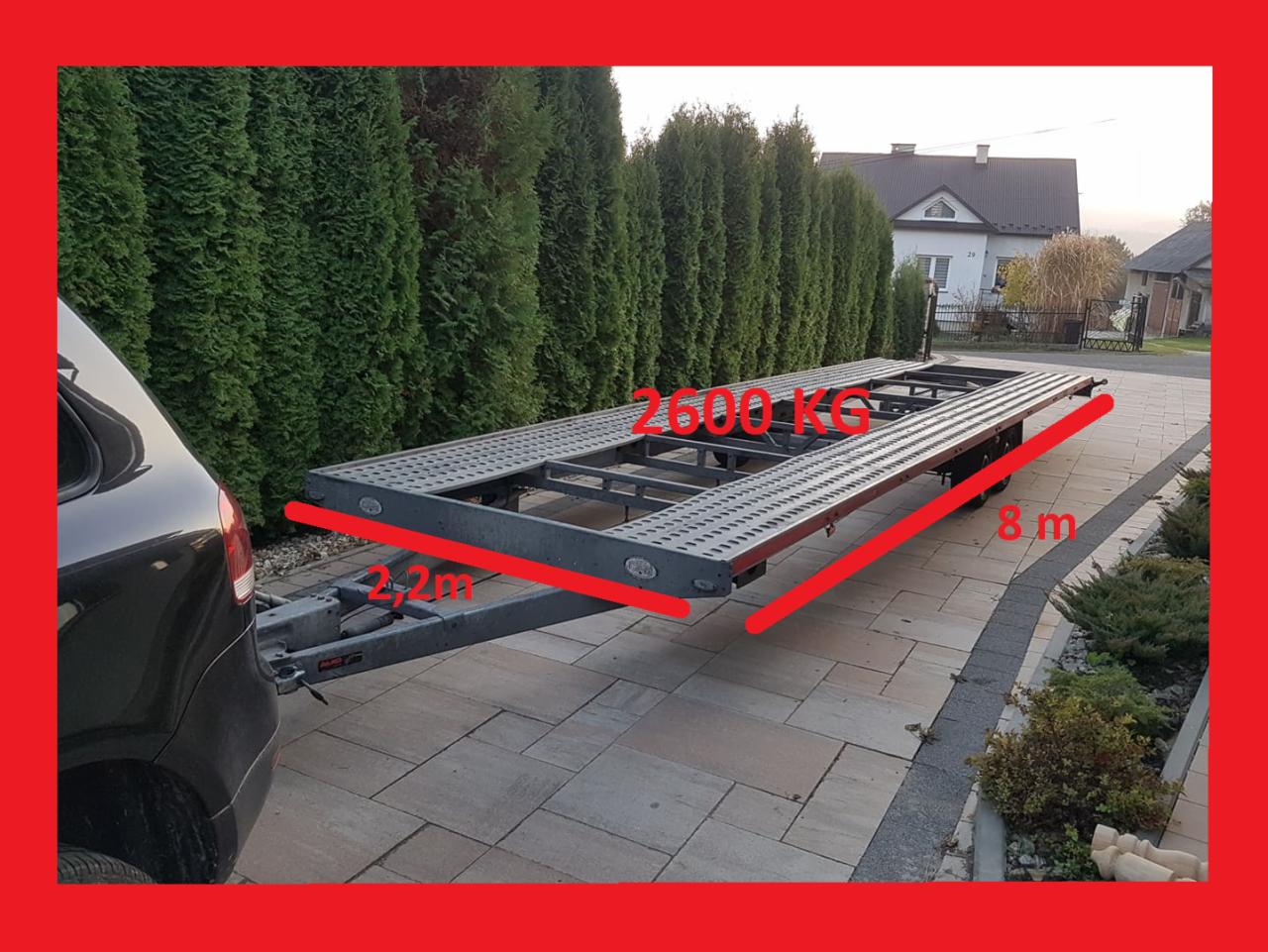 Czarny samochód osobowy z podpiętą, pustą lawetą o wymiarach 8m x 2.2m i ładowności 2600kg, zaparkowany na brukowanym podjeździe przed domem z tujami w tle.