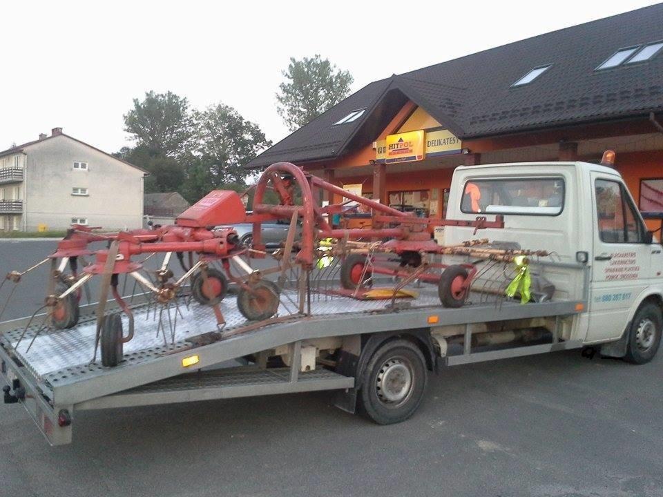 Czerwony przetrząsacz karuzelowy, maszyna rolnicza, przewożony na platformie białej lawety przed budynkiem z szyldem 'Delikatesy'.