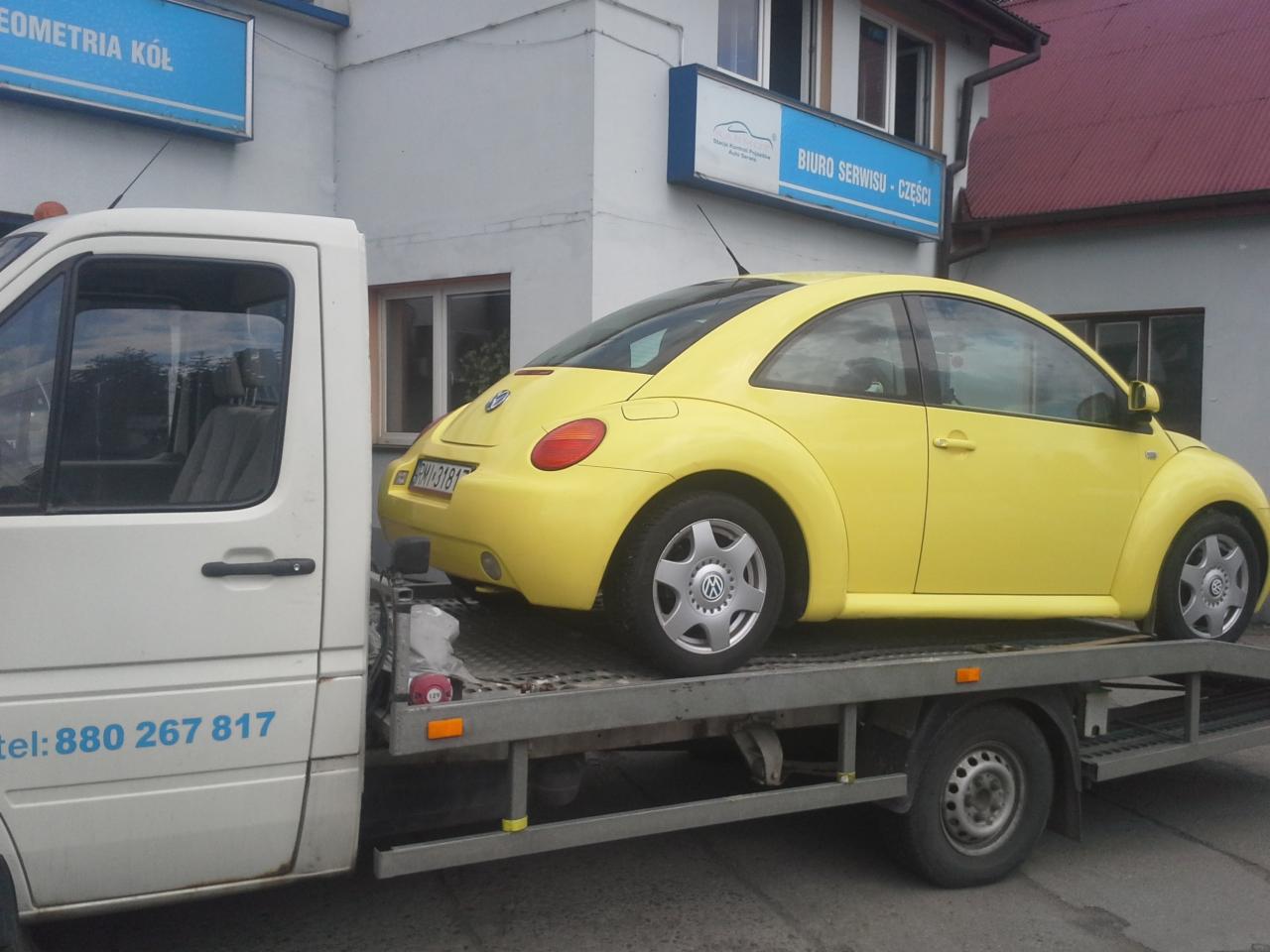 Żółty Volkswagen Beetle na platformie holowniczej, z numerem telefonu widocznym na kabinie pojazdu holującego, na tle budynku z szyldami warsztatu samochodowego.