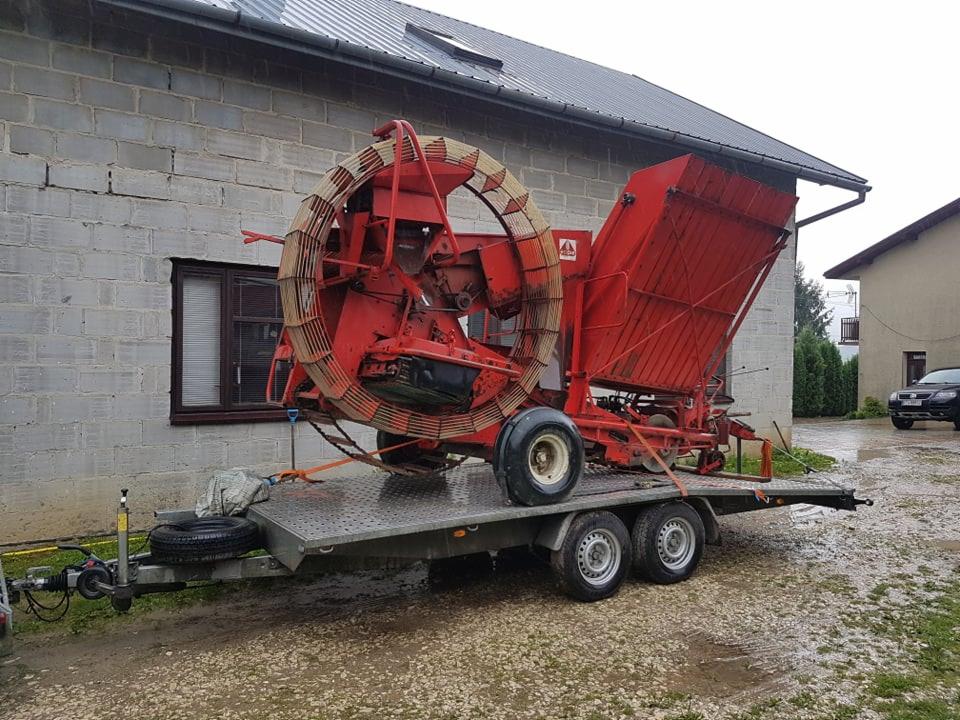 Czerwony kombajn rolniczy z okrągłym bębnem załadowany na niskopodwoziową przyczepę transportową, zabezpieczony pomarańczowymi pasami, na tle szarego budynku z cegły.