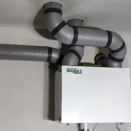 Biała jednostka centralna z szarymi rurami wentylacyjnymi przymocowanymi do sufitu, widoczne łączenia rur z czarną taśmą, ściany w stanie surowym, widoczna instalacja elektryczna w ścianie.