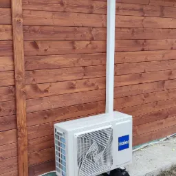 Zewnętrzna jednostka klimatyzacyjna marki Haier zamontowana na betonowej podstawie przy drewnianej ścianie budynku z brązową rynną.