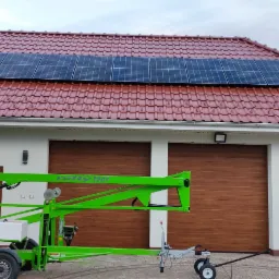 Panele słoneczne na czerwonym dachu domu jednorodzinnego, widoczne z poziomu podnośnika koszowego Nifty 120T, z otwartym garażem w tle.