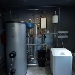 Zestaw urządzeń grzewczych w kotłowni: duży szary zbiornik, biała jednostka sterująca i liczne rury, zawory oraz czarne filtry na tle szarej ściany i podłogi z płytek imitujących marmur.