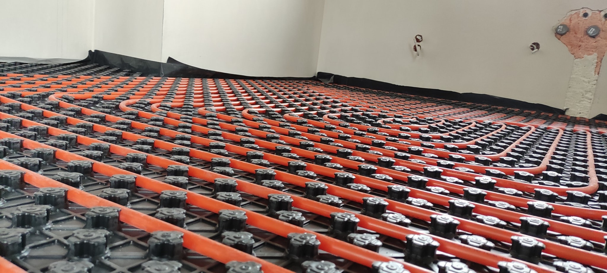 Pomarańczowe rury ogrzewania podłogowego ułożone w regularnych odstępach na czarnej macie montażowej, widoczne elementy mocujące, fragment ściany w tle z widocznymi puszkami instalacyjnymi.