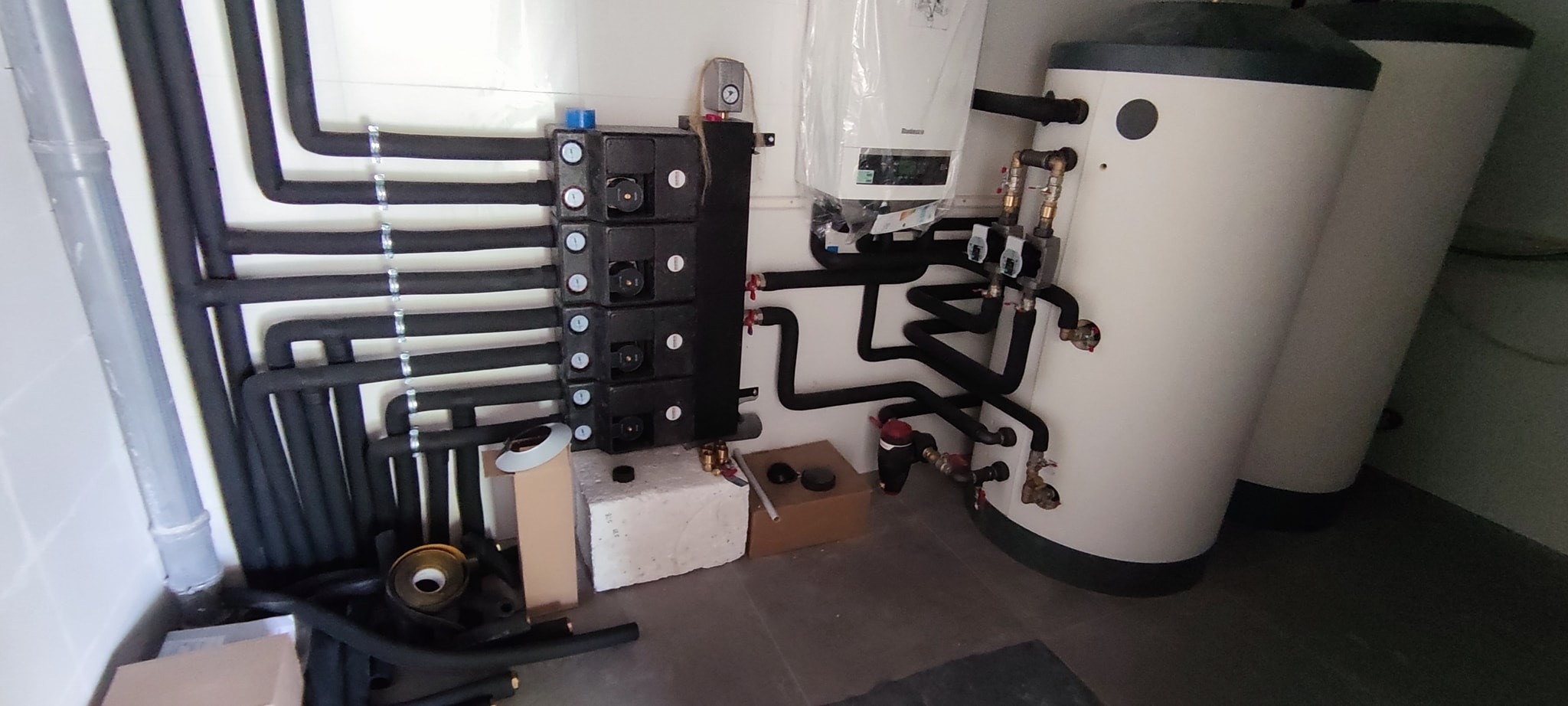 Kotłownia z dwoma białymi zbiornikami, czarnym rozdzielaczem, rurami w czarnej izolacji, pudełkami i akcesoriami hydraulicznymi na podłodze