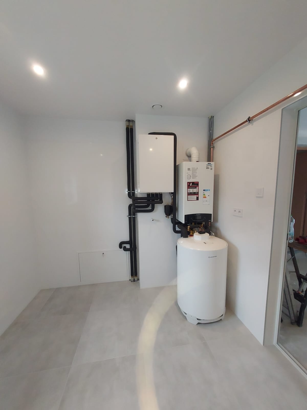 Biała kotłownia z zamontowanym dwufunkcyjnym kotłem gazowym Ariston, zasobnikiem ciepłej wody i czarnymi rurami instalacji grzewczej na tle białych ścian i szarej podłogi z płytek.