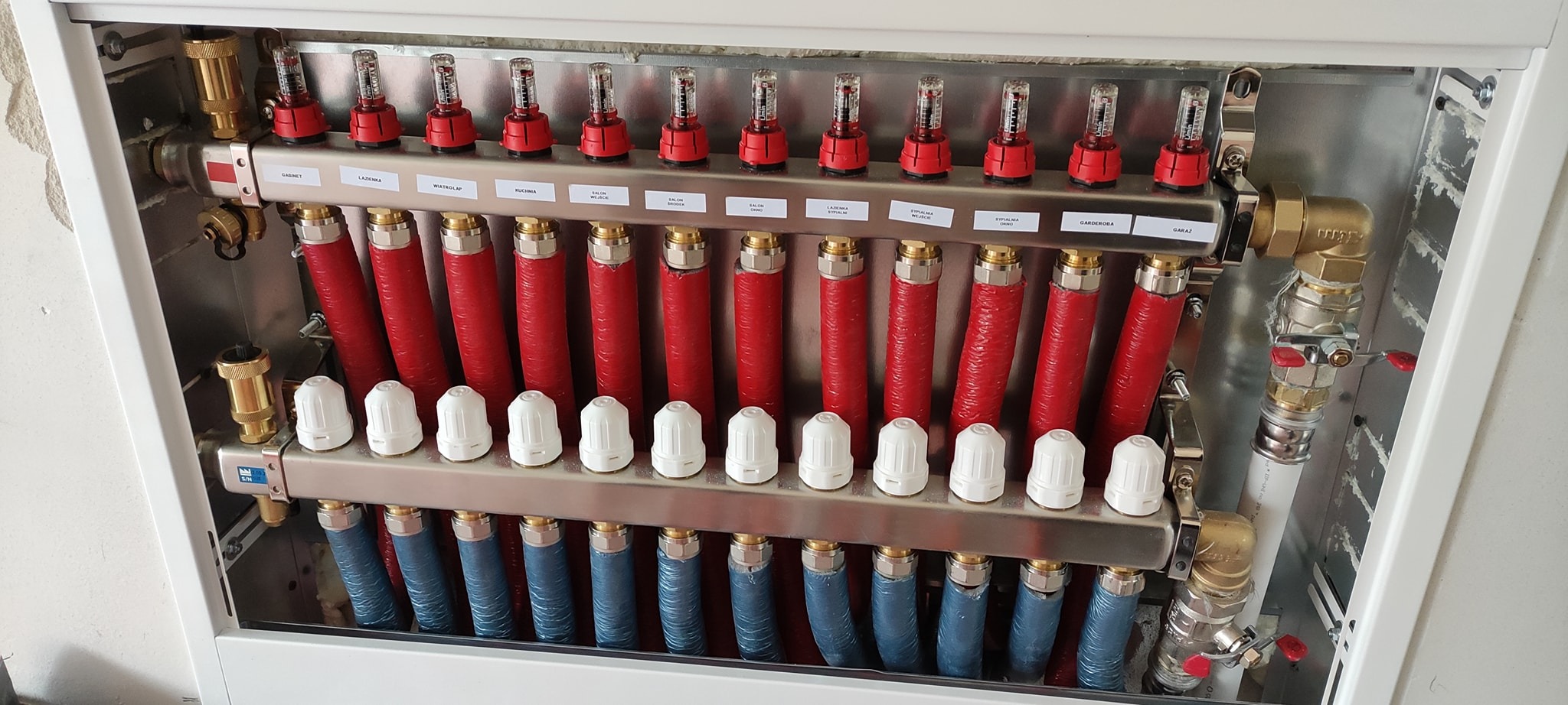 Rozdzielacz ogrzewania podłogowego w metalowej szafce, z czerwonymi i niebieskimi rurami, etykietami opisującymi pomieszczenia i przepływomierzami.