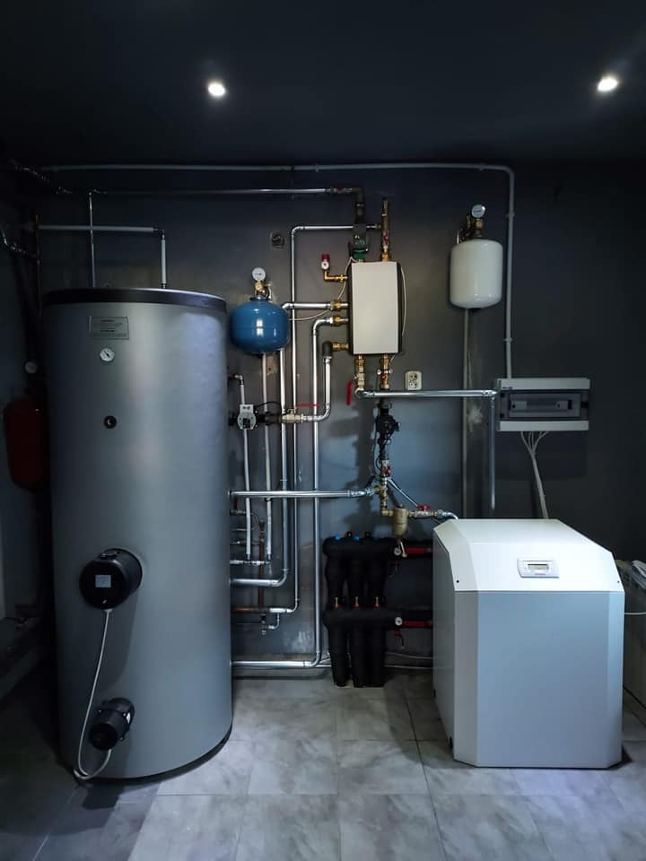 Zestaw urządzeń grzewczych w kotłowni: duży szary zbiornik, biała jednostka sterująca i liczne rury, zawory oraz czarne filtry na tle szarej ściany i podłogi z płytek imitujących marmur.