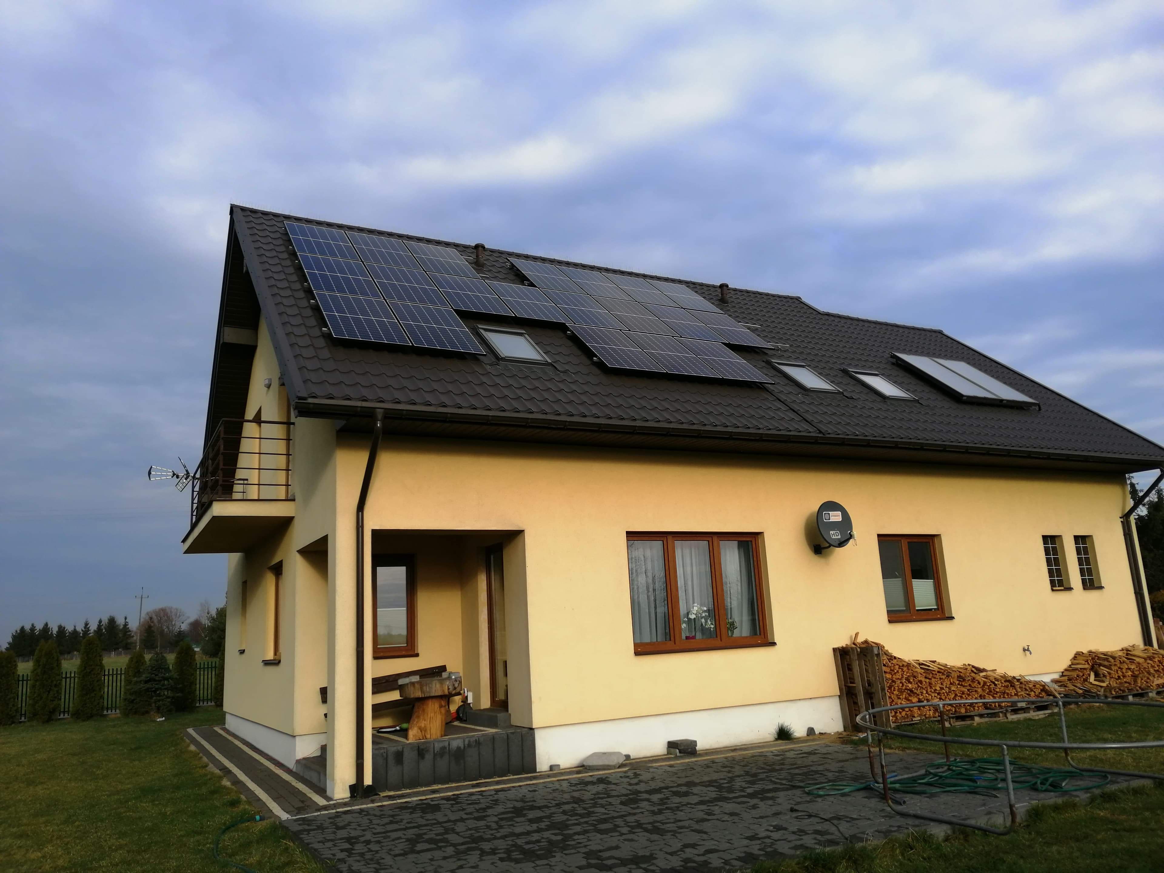 Żółty dom jednorodzinny z ciemnobrązowym dachem pokrytym panelami fotowoltaicznymi i oknami dachowymi, widok z zewnątrz na tle pochmurnego nieba.