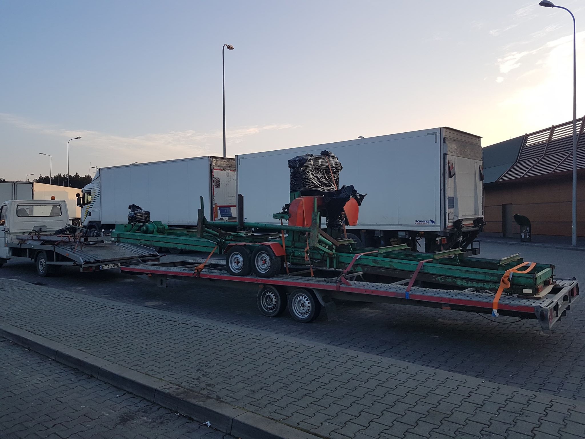 Laweta z zieloną konstrukcją i ładunkiem zabezpieczonym czarną folią, ciągnięta przez małą lawetę, na parkingu z białymi ciężarówkami w tle, wczesny wieczór.