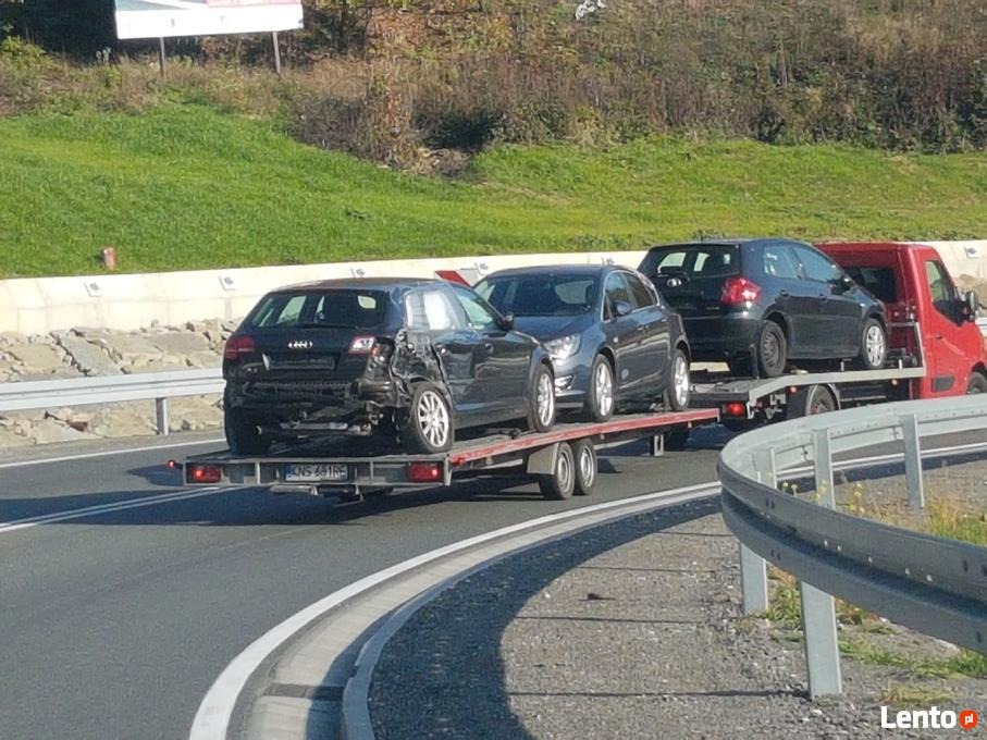 Laweta przewozi trzy samochody, w tym jeden uszkodzony z tyłu, na autostradzie w słoneczny dzień.