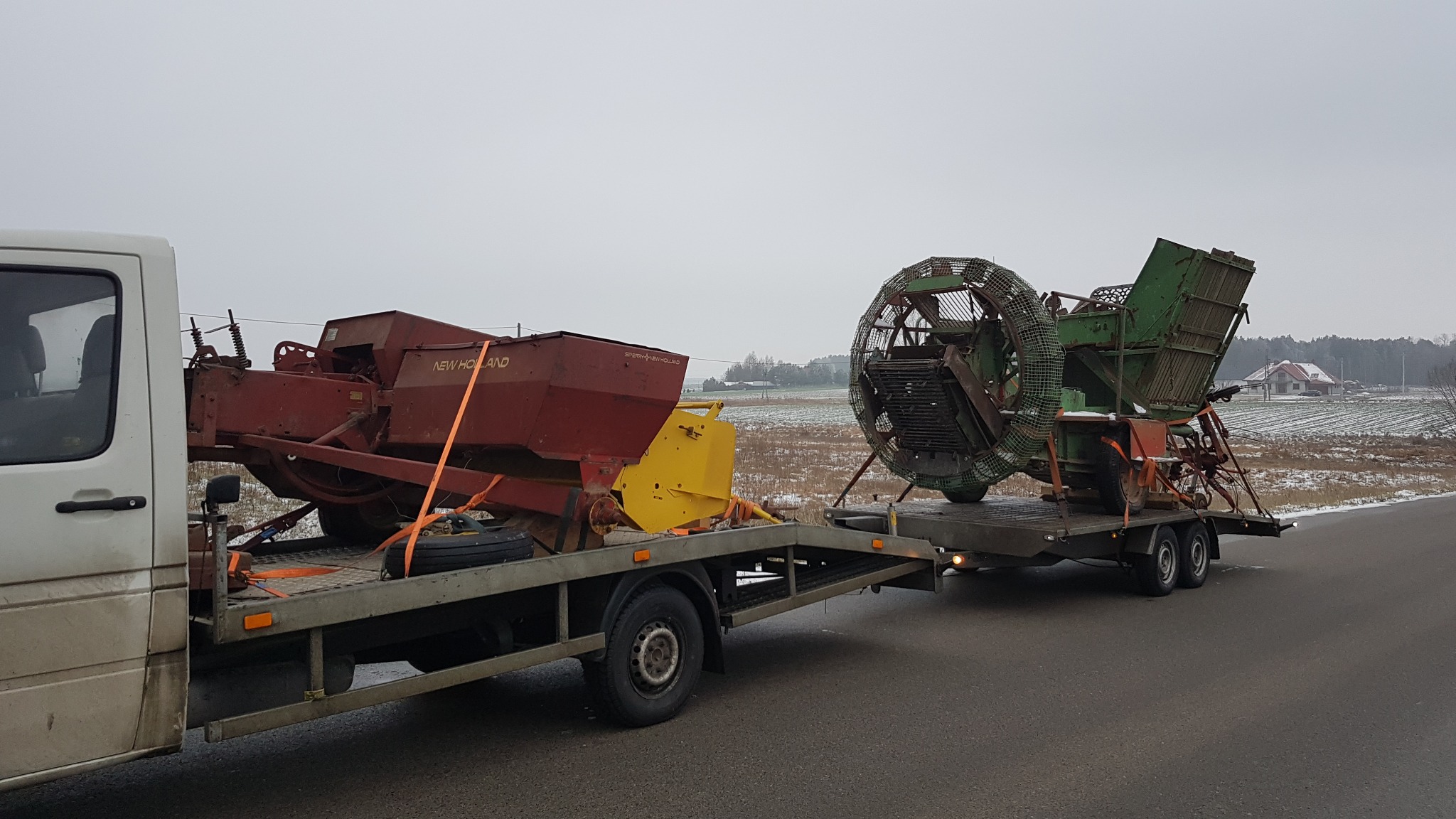 Laweta przewozi używane maszyny rolnicze: kombajn ziemniaczany i siewnik New Holland, zabezpieczone pomarańczowymi pasami, na tle zimowego krajobrazu z polem i domami.