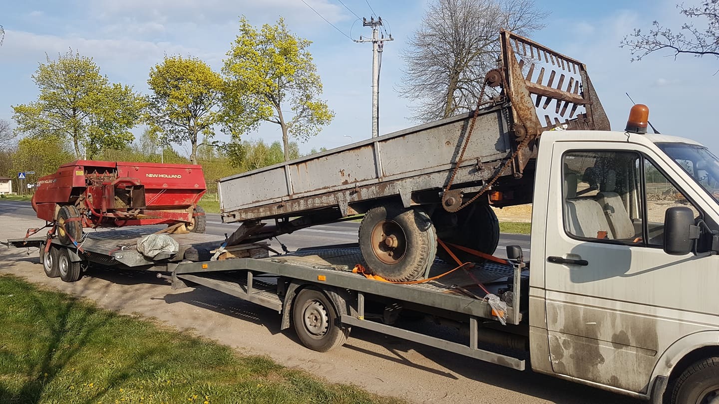 Biała laweta przewozi przyczepę z prasą rolniczą New Holland i przyczepę rolniczą na tle zieleni.