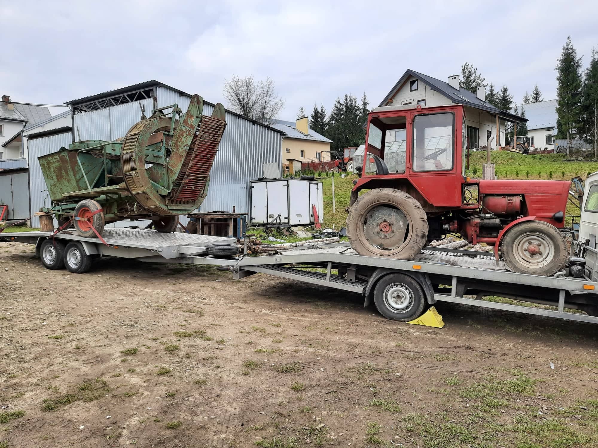 Czerwony traktor z zieloną maszyną rolniczą załadowane na srebrną lawetę na tle wiejskich budynków i pochmurnego nieba.