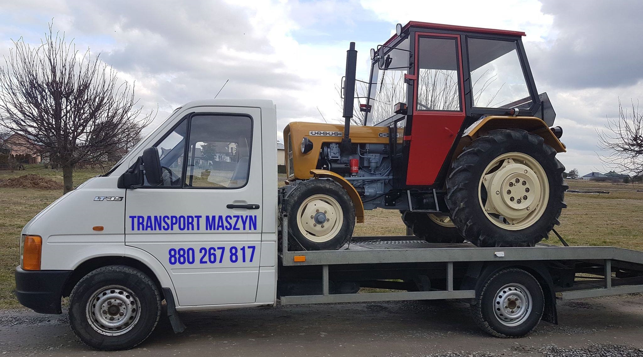 Biały samochód dostawczy z niebieskim napisem 'TRANSPORT MASZYN 880 267 817' przewozi na platformie ciągnik rolniczy Ursus C-330 w kolorach żółtym i czerwonym.