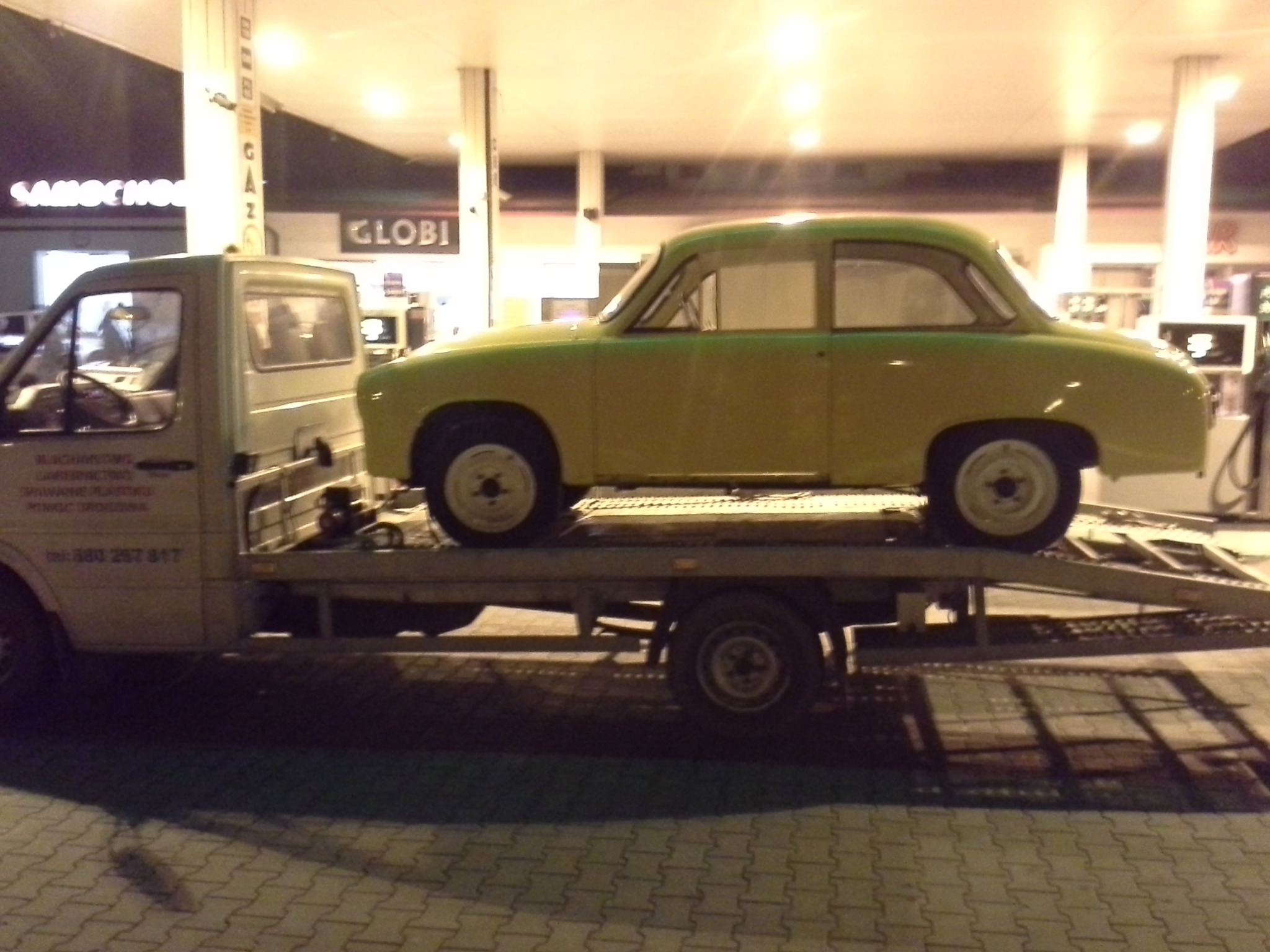 Żółty samochód osobowy, typu Syrena, przewożony na platformie lawety na tle stacji benzynowej oświetlonej nocą.